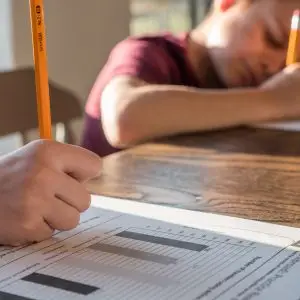 Crop unrecognizable schoolkid with pencil in hand writing on paper sheet with diagrams while sitting at table with brother and doing homework
